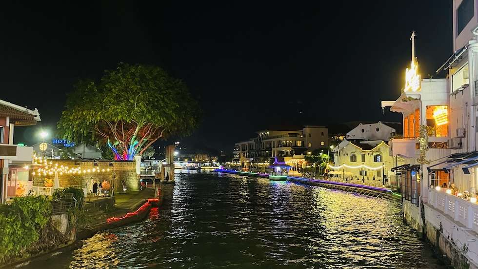 View down Melaka river at night. Colourful lights are strung on either side of the bank.