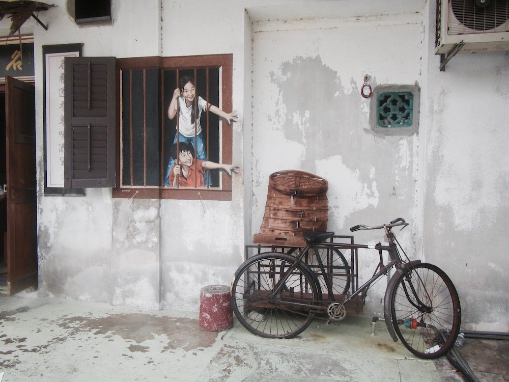 Travel guide 'Looking Back to Penang Street Art and Around the Island' on sustainable travel blog Teja on the Horizon | Two children reaching out for pau from a basket on a bicycle | Penang street art