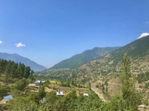 View down the mountain valley summer countryside in Dachigam National Park, Kashmir. The sky is clear and blue, and the hillsides are partially dotted by trees.