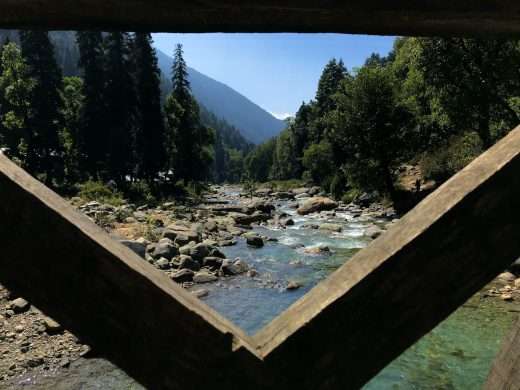 View through a wooden bridge frame along a mountain creek in Kashmir. River rocks line the banks and tall conifer trees flank sides of the narrow creek. The bridge frame forms a V in the foreground.