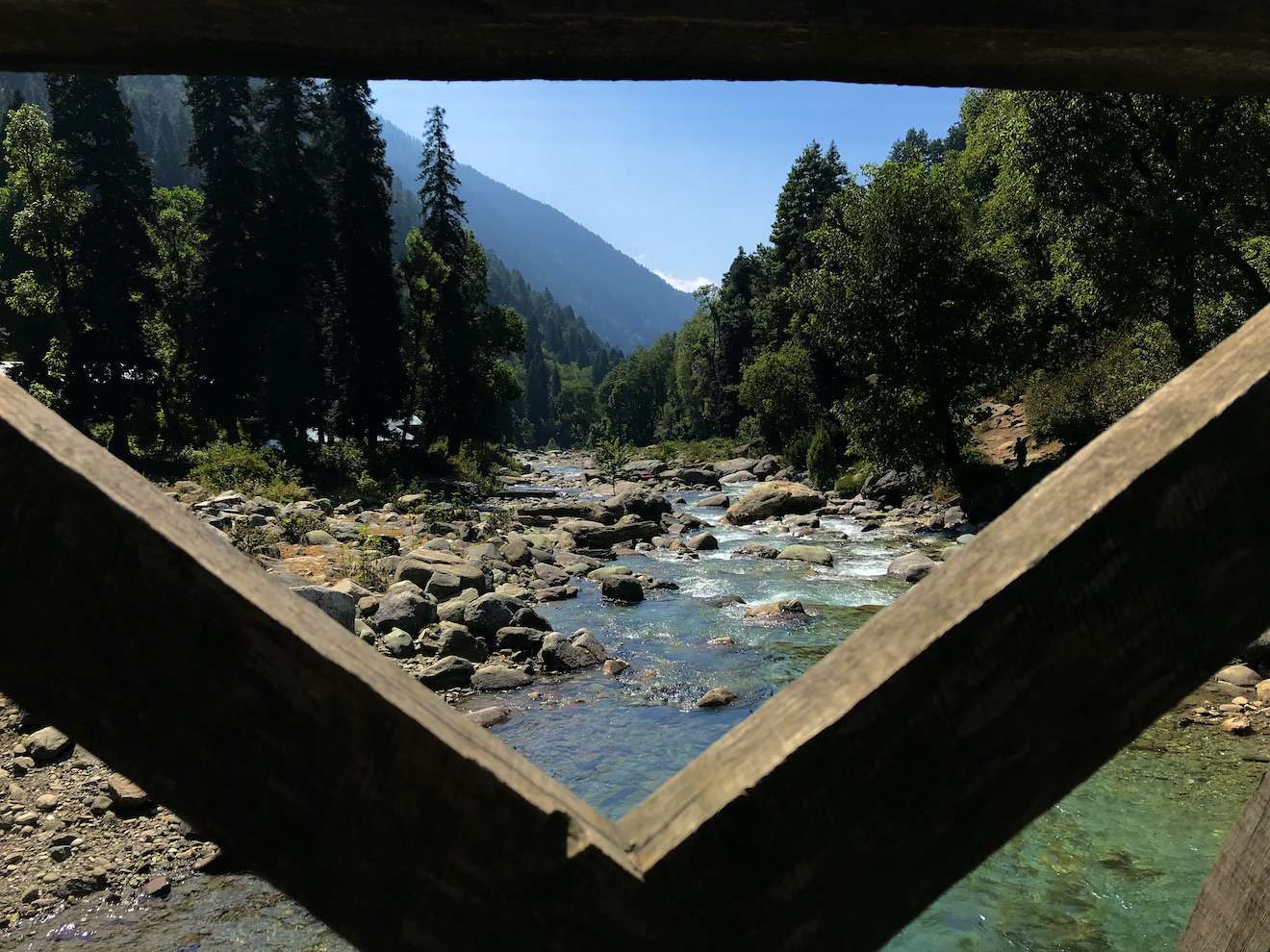 View through a wooden bridge frame along a mountain creek in Kashmir. River rocks line the banks and tall conifer trees flank sides of the narrow creek. The bridge frame forms a V in the foreground.