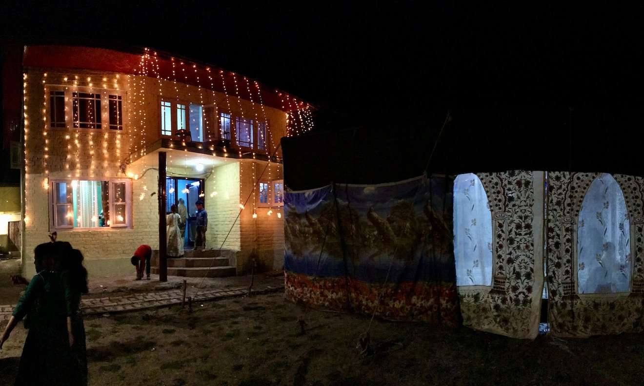 Village compound in Srinagar decked out for a wedding. The house is hung with warm orange fairy lights, and a traditional eating tent featuring Kashmir's hamdan embroidery is set up nearby.