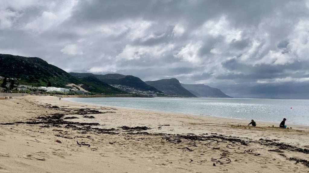 Two children making sand castles on the beach near Simon's Town railway station. Kelp lie strewen on the yellowish sand where the high tide mark had been. The steep headlands of the Cape peninsula coast extend in series in the background. The sky is heavily overcast.